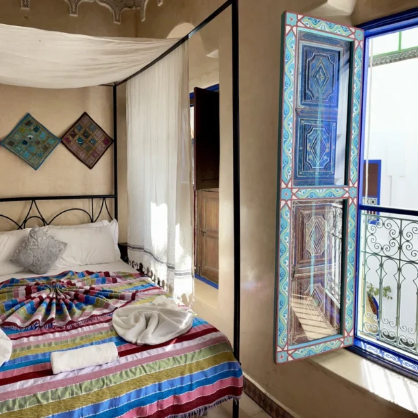 Cozy riad room featuring a decorated bed, white canopy, and open painted shutters overlooking the courtyard.