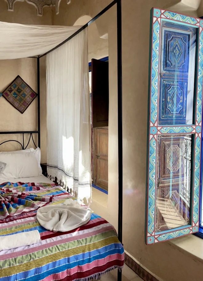 Cozy riad room featuring a decorated bed, white canopy, and open painted shutters overlooking the courtyard.