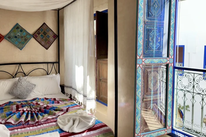Cozy riad room featuring a decorated bed, white canopy, and open painted shutters overlooking the courtyard.